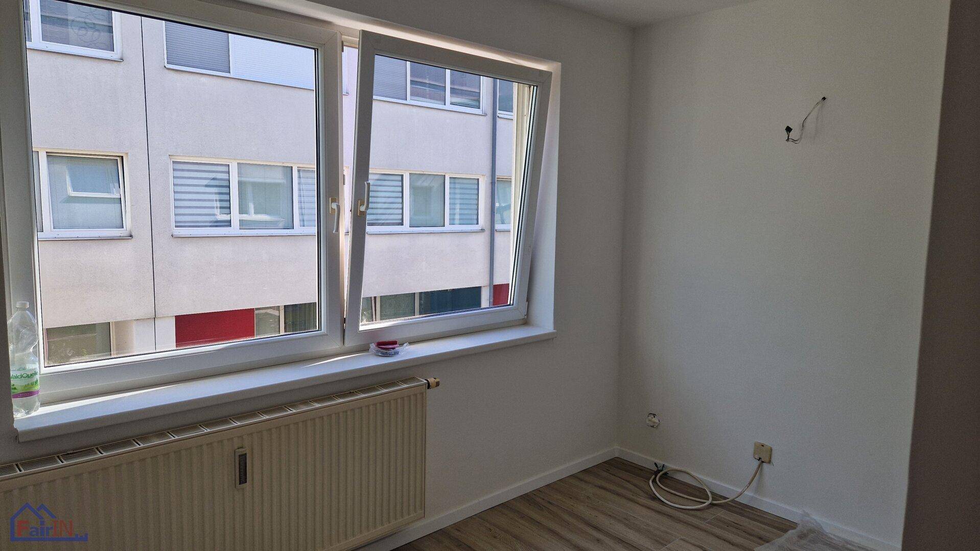 Leeres Schlafzimmer mit Laminatboden und einem Fenster, das den Blick auf ein gegenüberliegendes Gebäude freigibt.