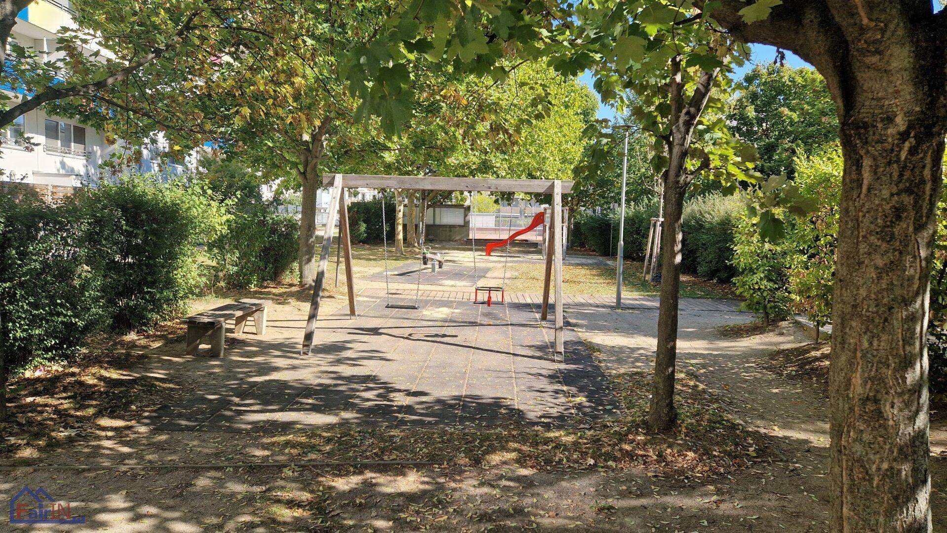Grüner Spielplatz mit Schaukeln und Rutsche, umgeben von Bäumen und Sträuchern.