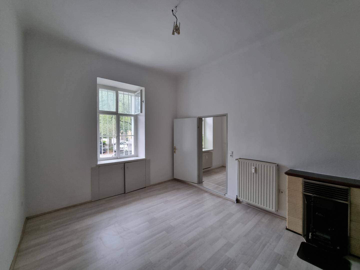 Geräumiges Zimmer mit Laminatboden, Fenster und einem traditionellen Kachelofen als Heizquelle.