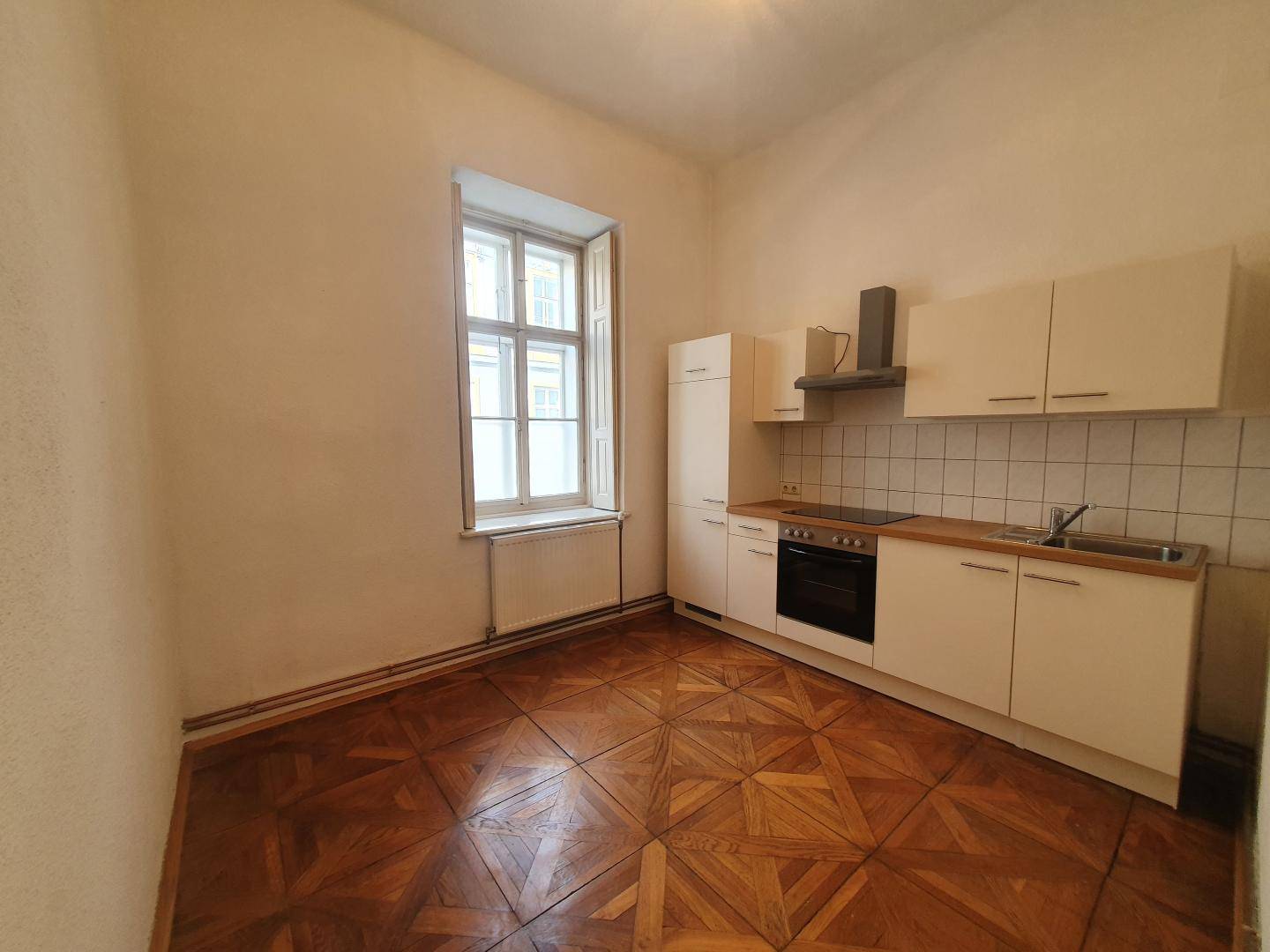 Funktionale Küche mit weißen Schränken, Herd und Spüle, ausgestattet mit Parkettboden und Fenster.