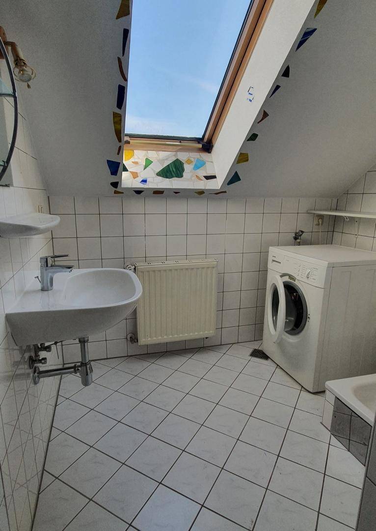 Badezimmer im Dachgeschoss mit Waschmaschine, Fliesenboden und farbigem Dachfenster.