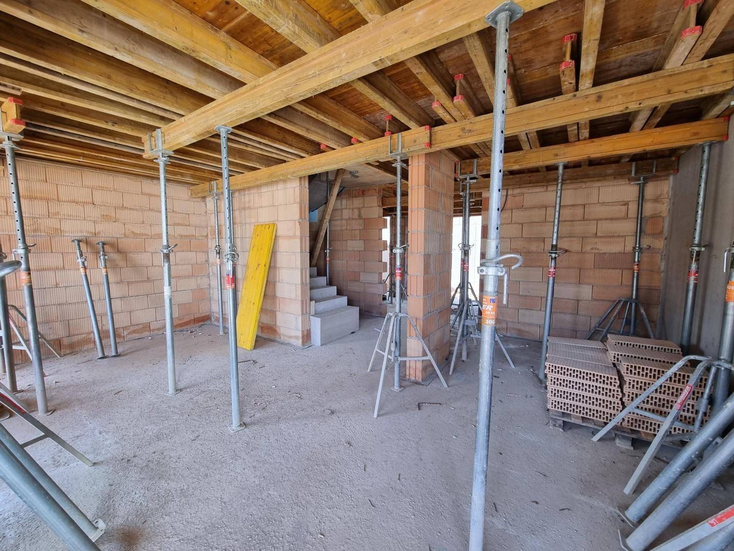 Innenansicht eines Rohbaus mit Ziegelwänden, Betonboden und Deckenstützen aus Holz und Metall.