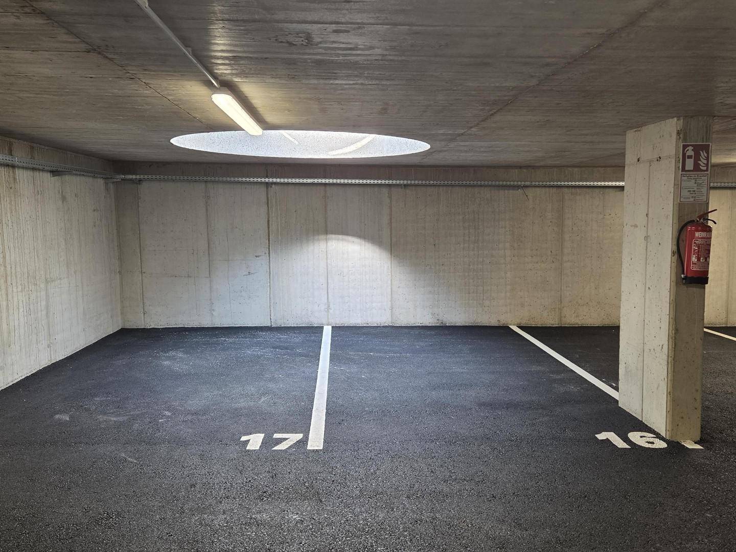 Tiefgaragenstellplätze mit Betonwänden und Decke, beleuchtet von Leuchtstoffröhren und einer runden Öffnung.