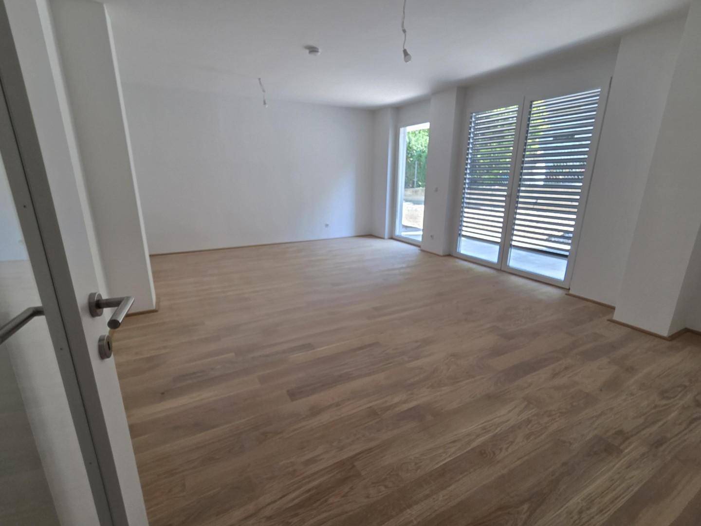 Geräumiges Wohnzimmer mit Holzboden und großen Fenstern, die viel Tageslicht hereinlassen.