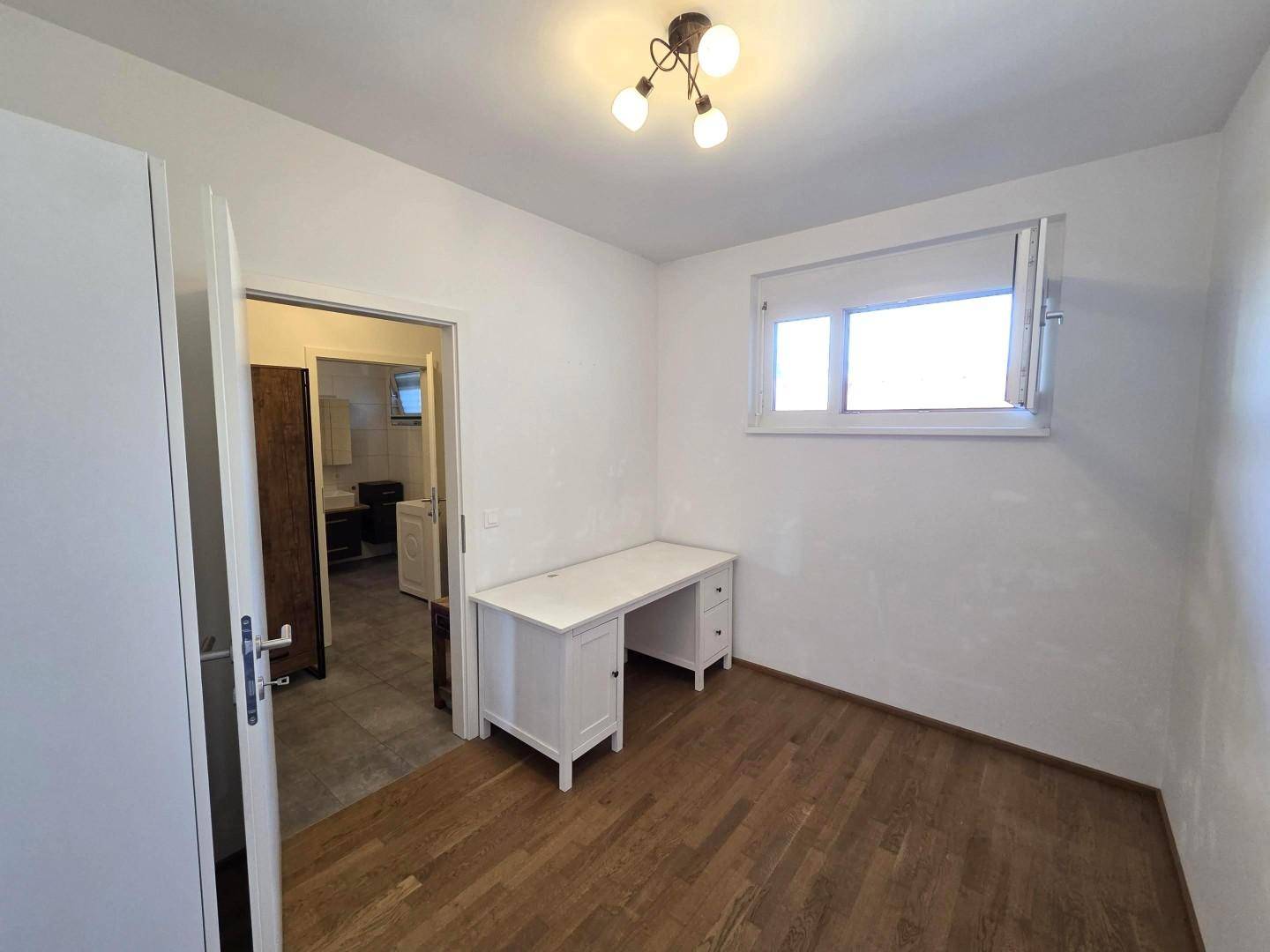 Helles Arbeitszimmer mit Schreibtisch, Holzboden und Fenster, mit Zugang zu einem Nebenraum.