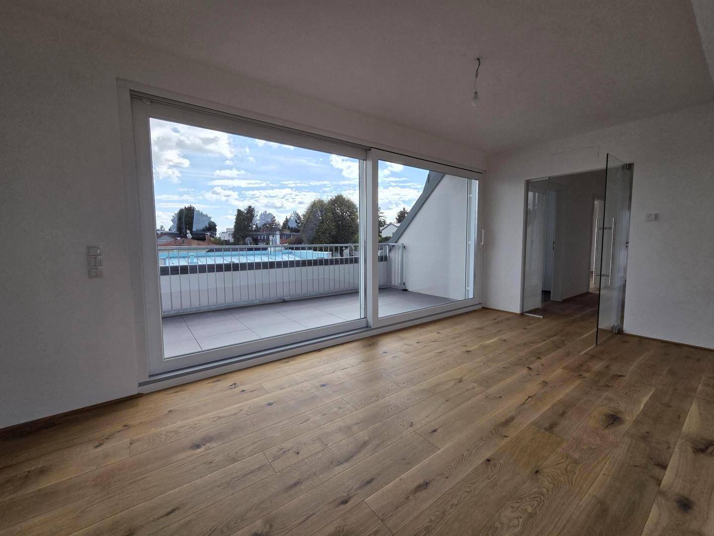 Geräumiger, heller Raum mit Holzboden und breiter Schiebetür, die zu einem Balkon führt.