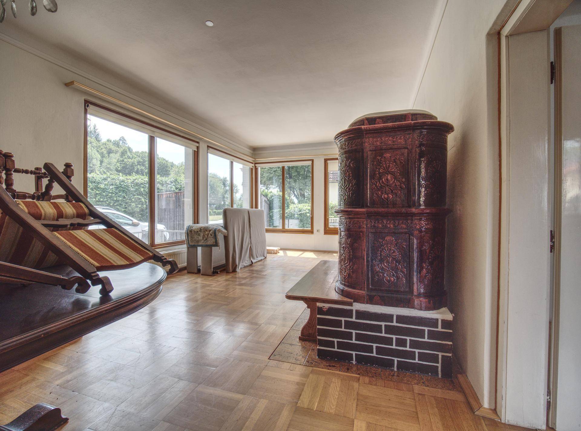 Helles Wohnzimmer mit Kachelofen, Parkettboden und großen Fenstern mit Blick ins Grüne.