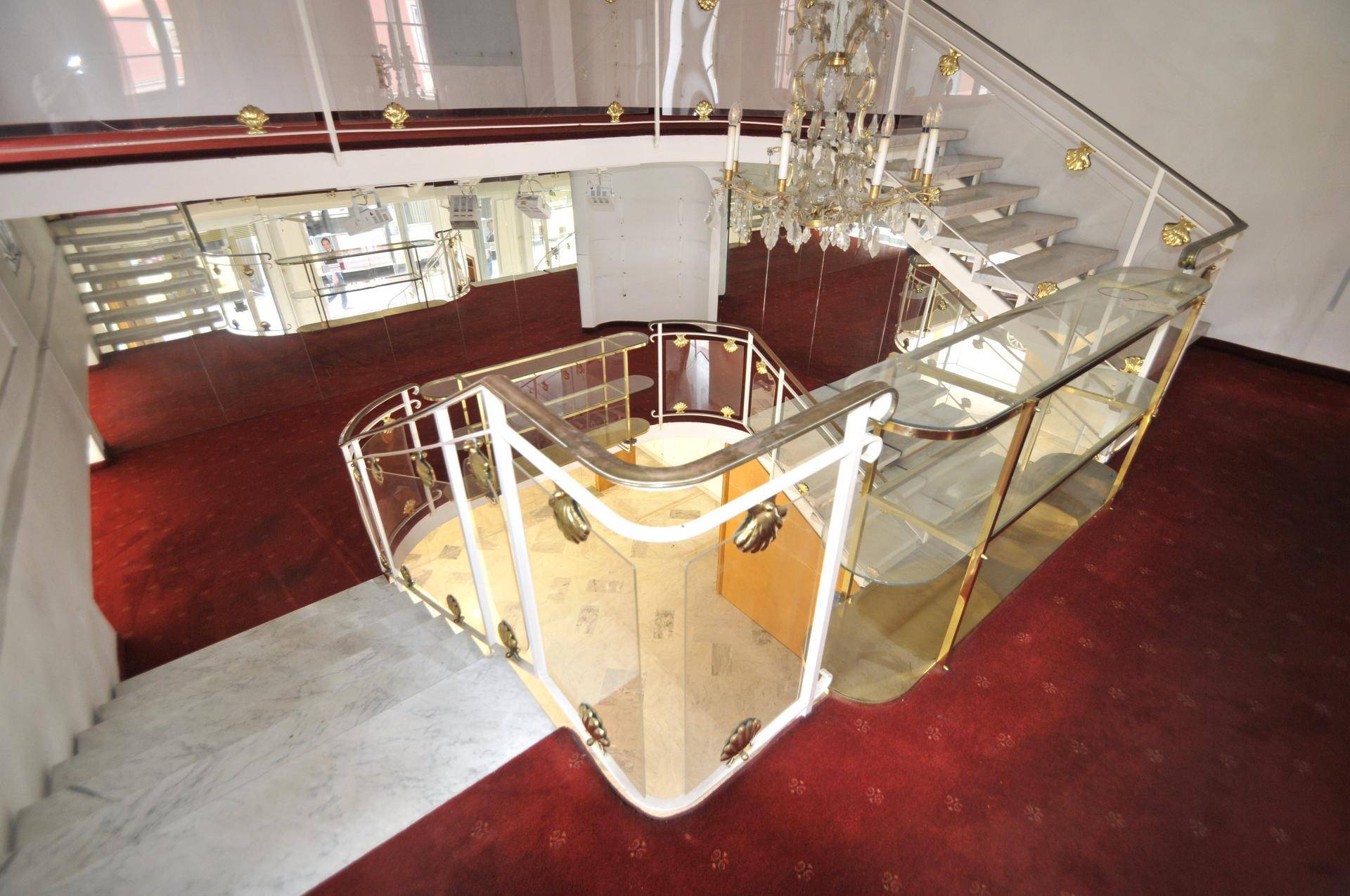 Blick von oben auf die geschwungene Treppe mit Glasgeländer und rotem Teppichboden im unteren Bereich.