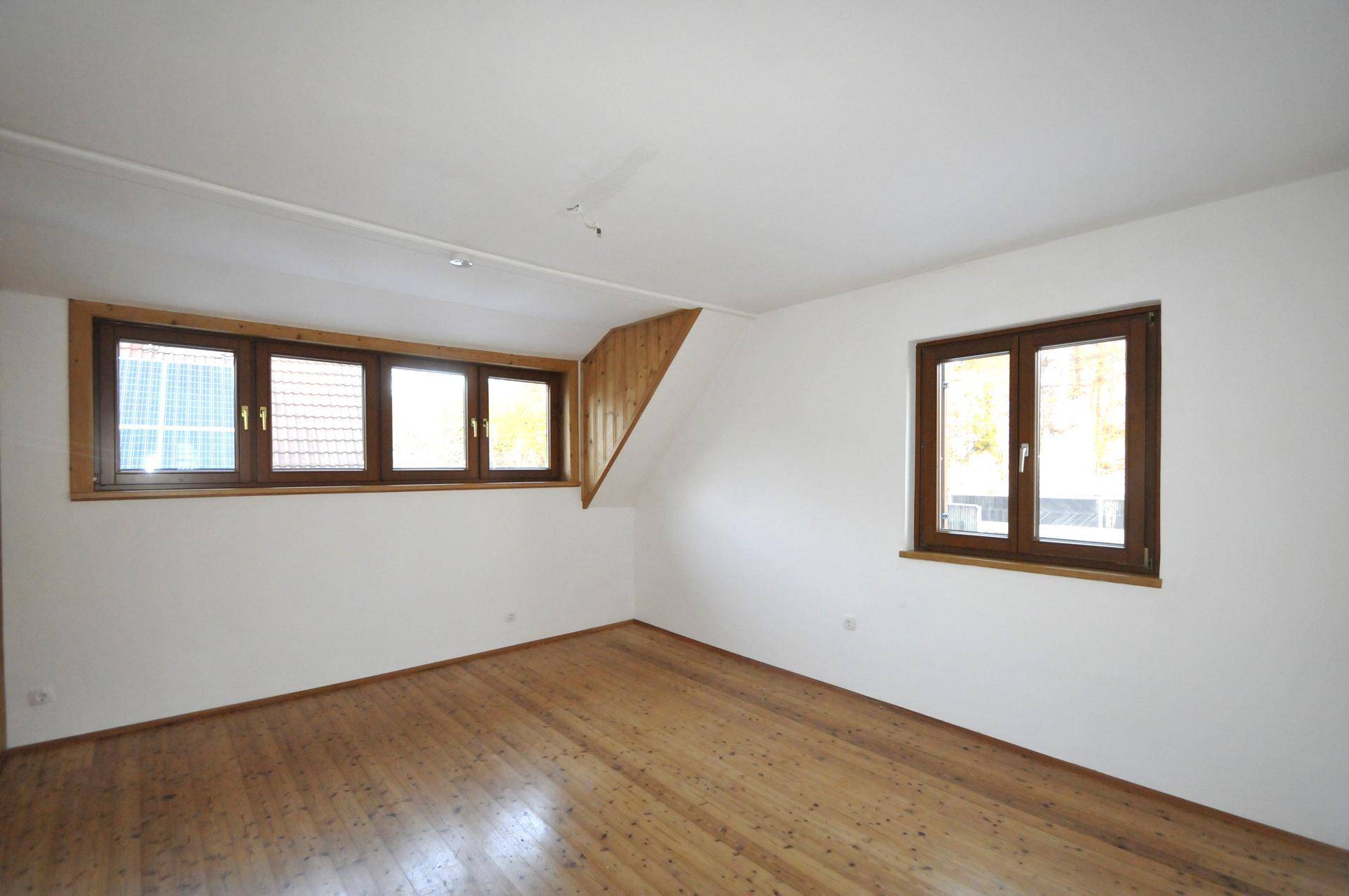 Heller Raum mit Holzboden, weißen Wänden und mehreren Fenstern, teilweise mit Dachschrägen.
