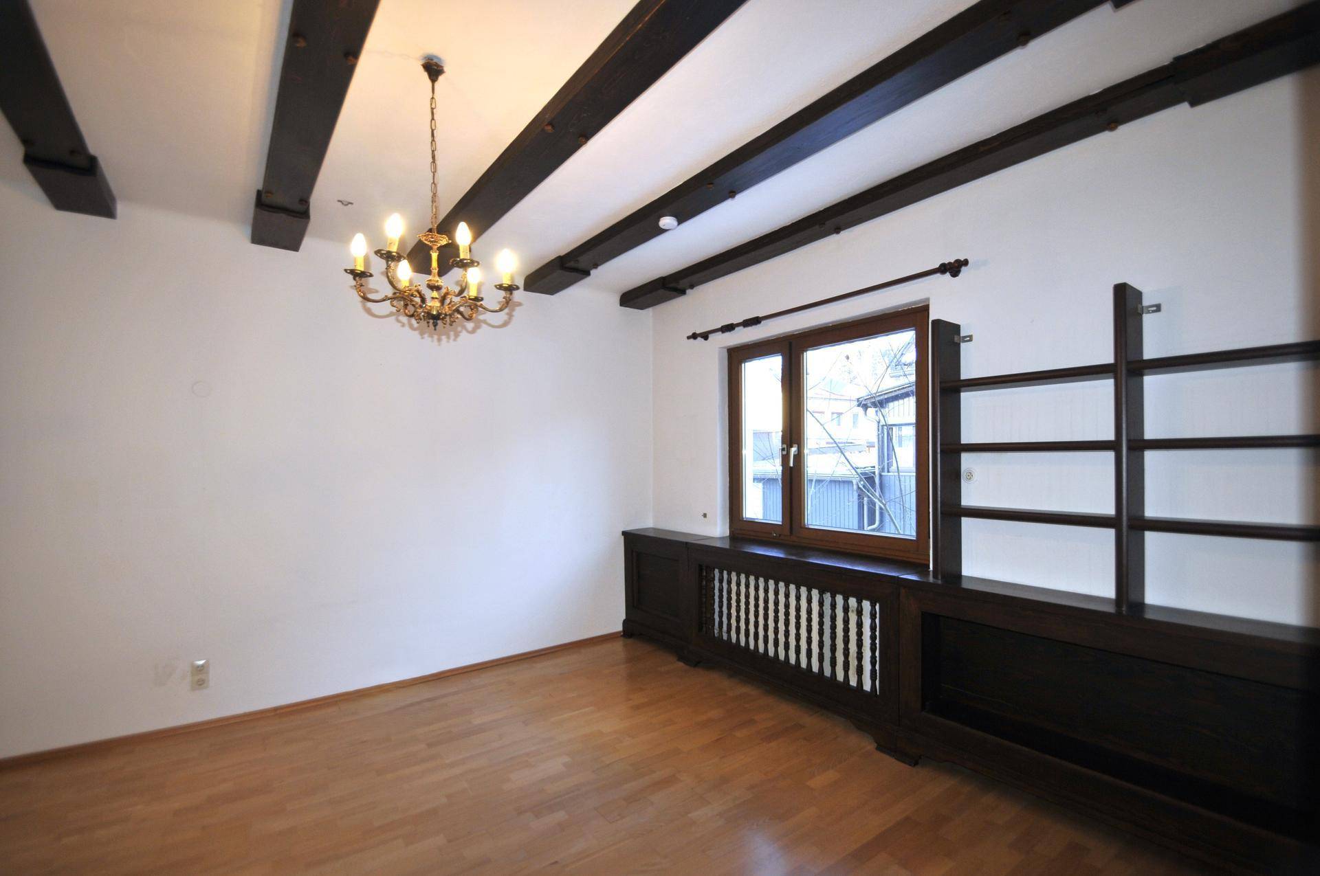 Gemütlicher Raum mit Holzboden, sichtbaren Deckenbalken und einem Fenster mit dekorativer Heizkörperverkleidung.