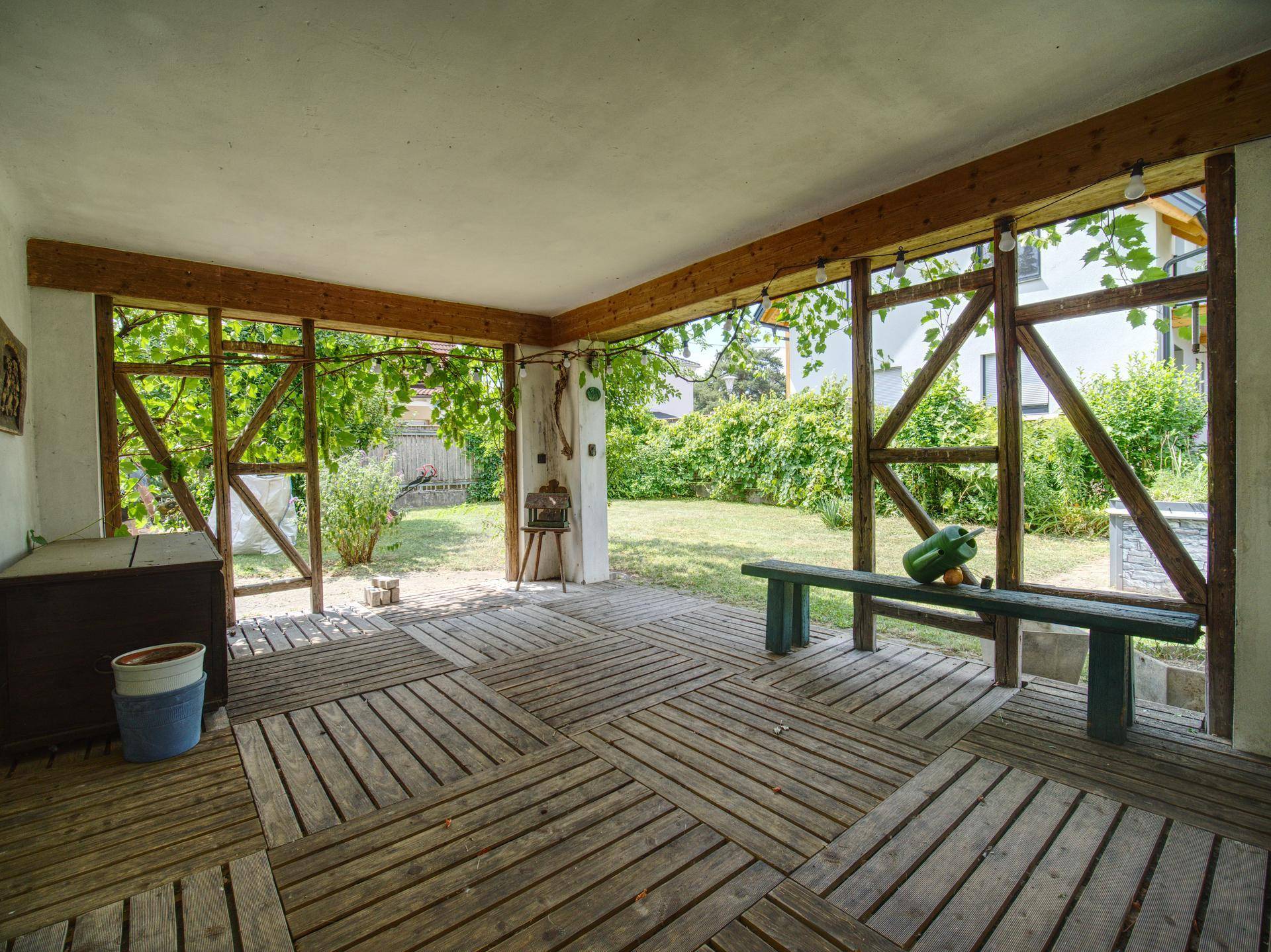 Gemütlicher überdachter Terrassenbereich mit Holzboden und Sitzbank, umgeben von viel Grün.