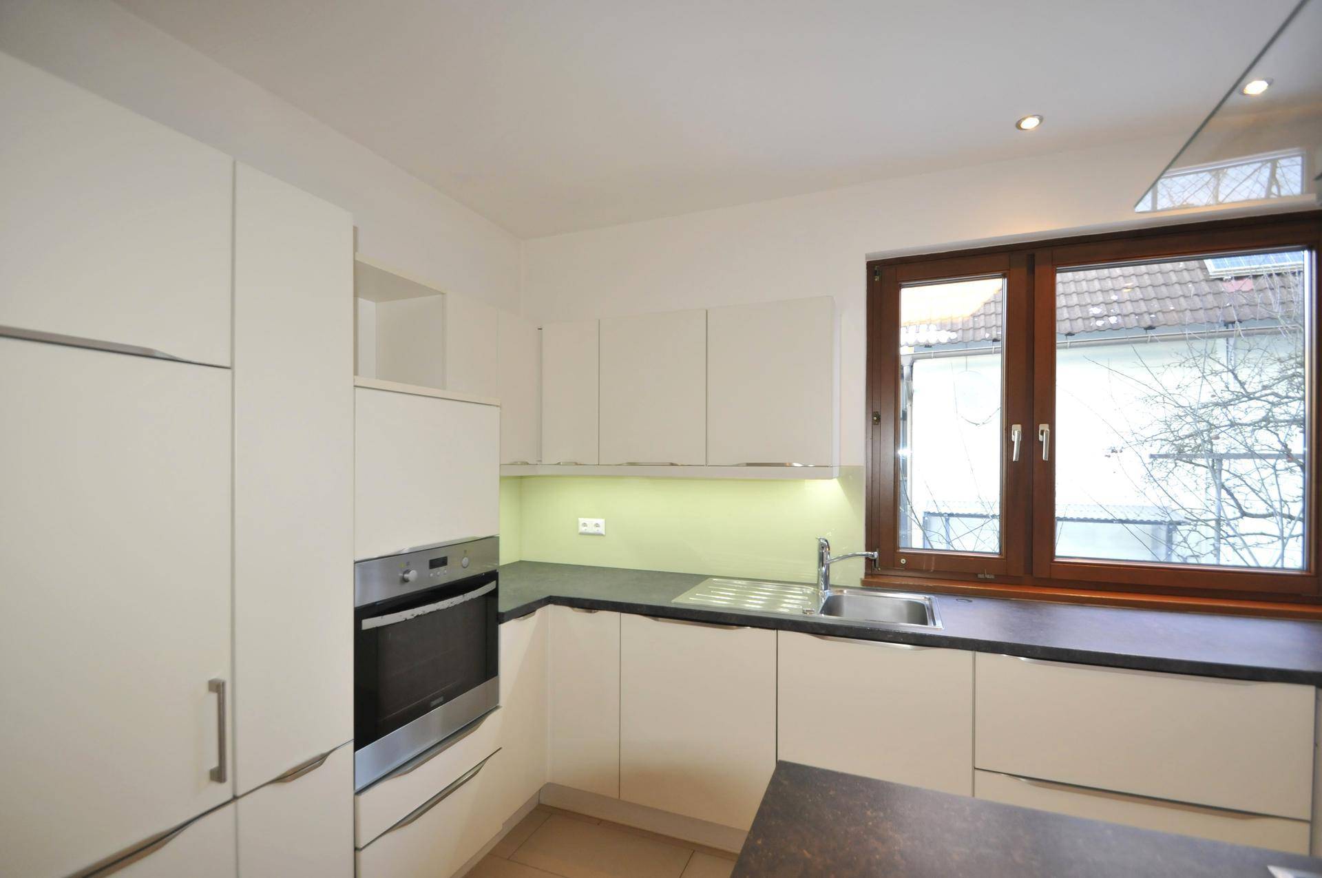 Moderne Küche mit weißen Schränken, dunkler Arbeitsplatte, Einbauofen und großem Fenster.