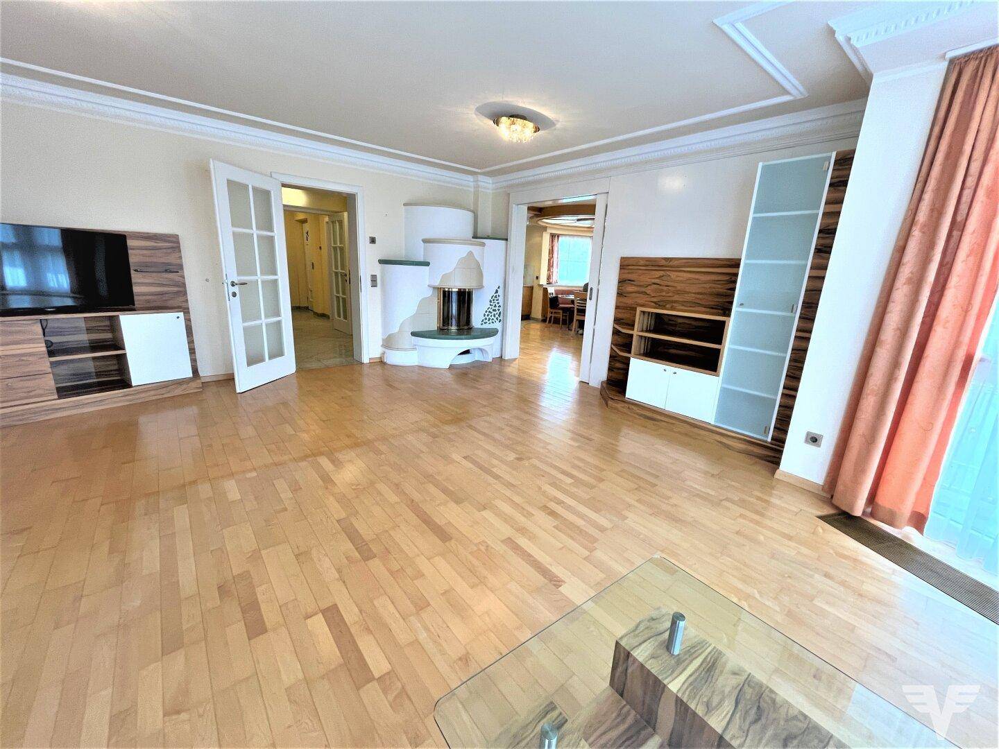 Helles Wohnzimmer mit Parkettboden, Kachelofen und Zugang zu weiteren Räumen.