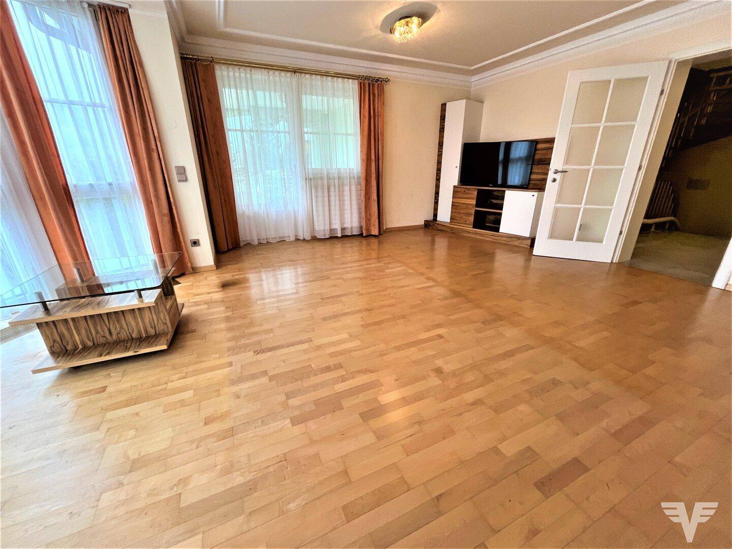 Geräumiges Wohnzimmer mit großen Fenstern, Parkettboden und viel Tageslicht.