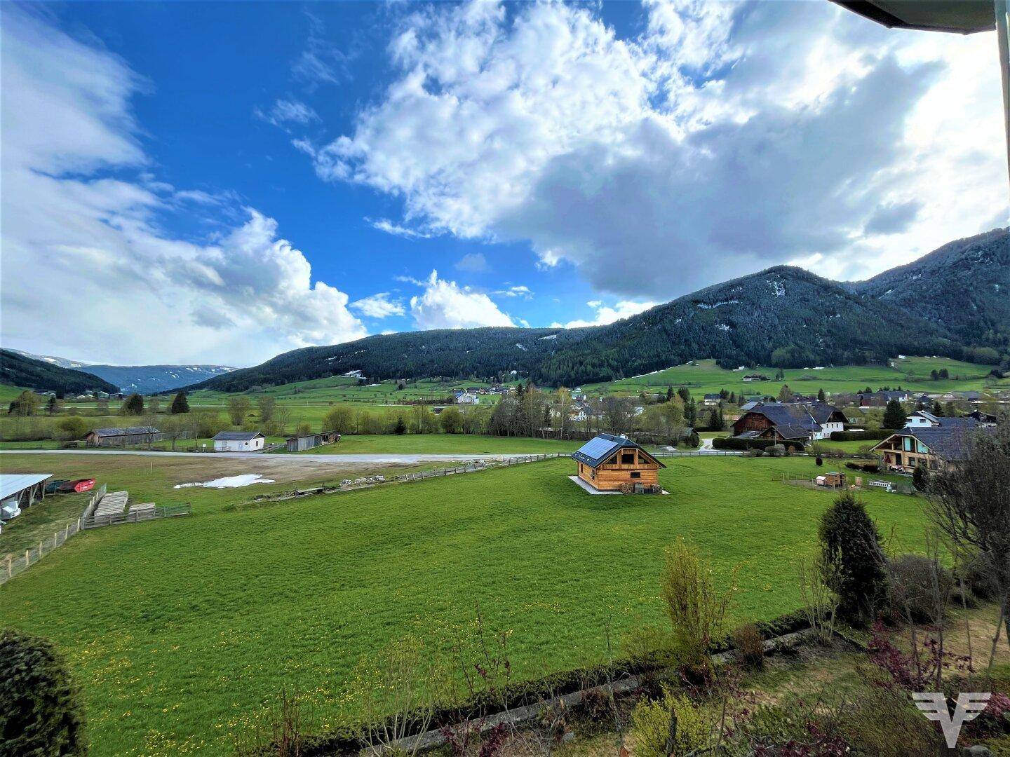 Weitläufiger Blick über grüne Felder und eine ländliche Umgebung mit Bergen im Hintergrund.