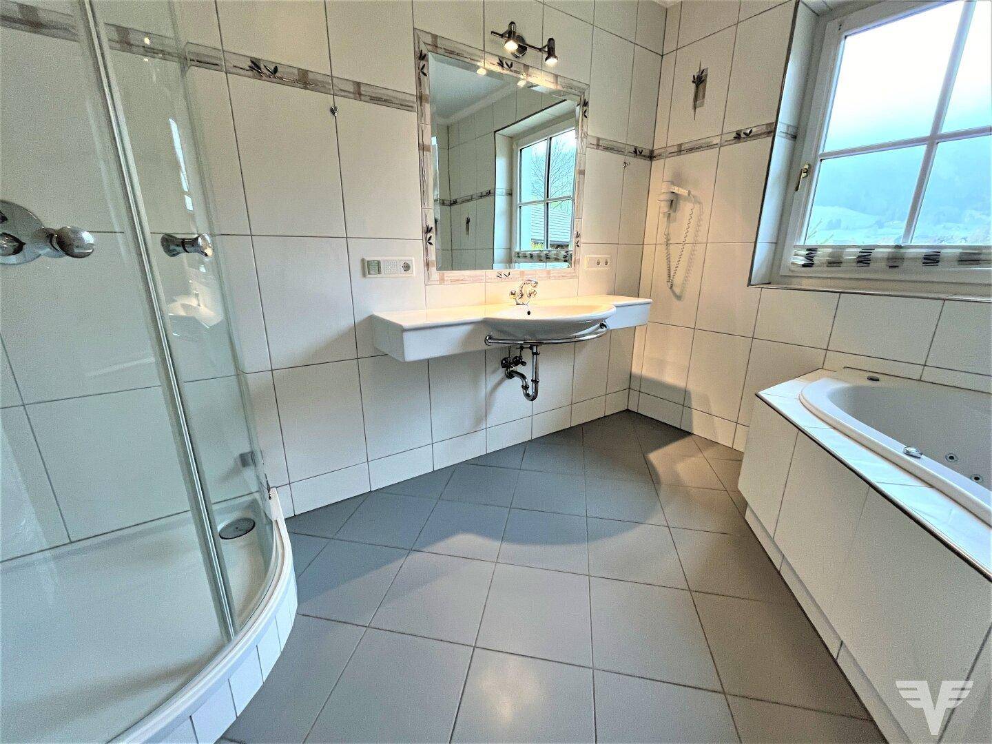 Badezimmer mit weißen Fliesen, Dusche, Eckbadewanne und Fenster für Tageslicht.