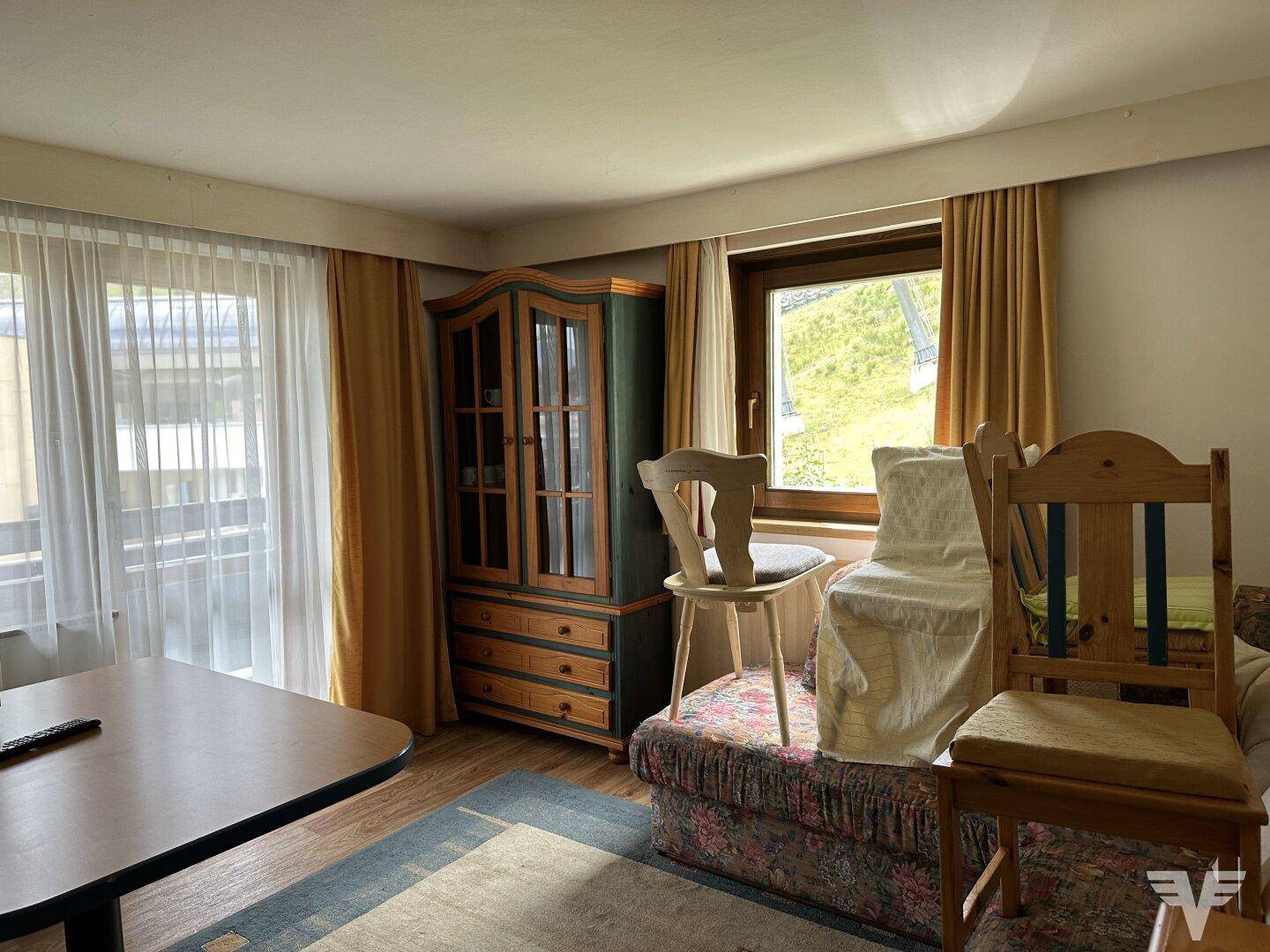 Gemütlicher Wohn- und Essbereich mit traditionellen Holzmöbeln, Teppich und Blick ins Grüne durch die Fenster.