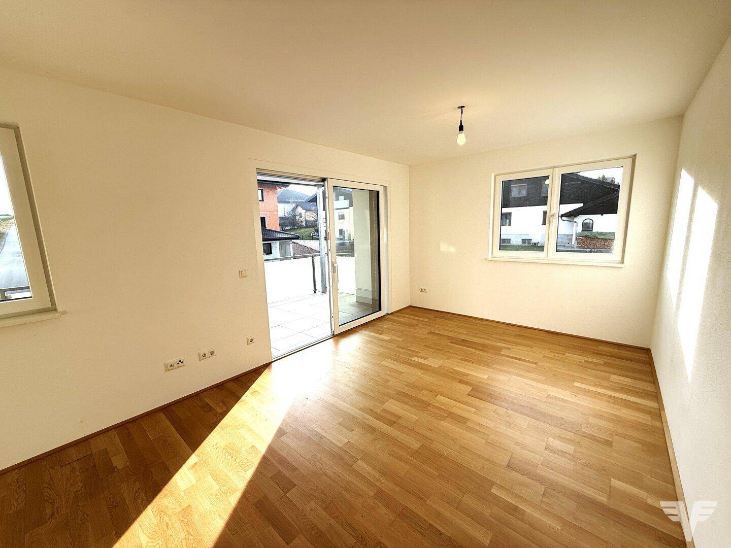 Heller Wohnraum mit Holzboden und Blick auf die Umgebung durch große Fenster und Balkontür.