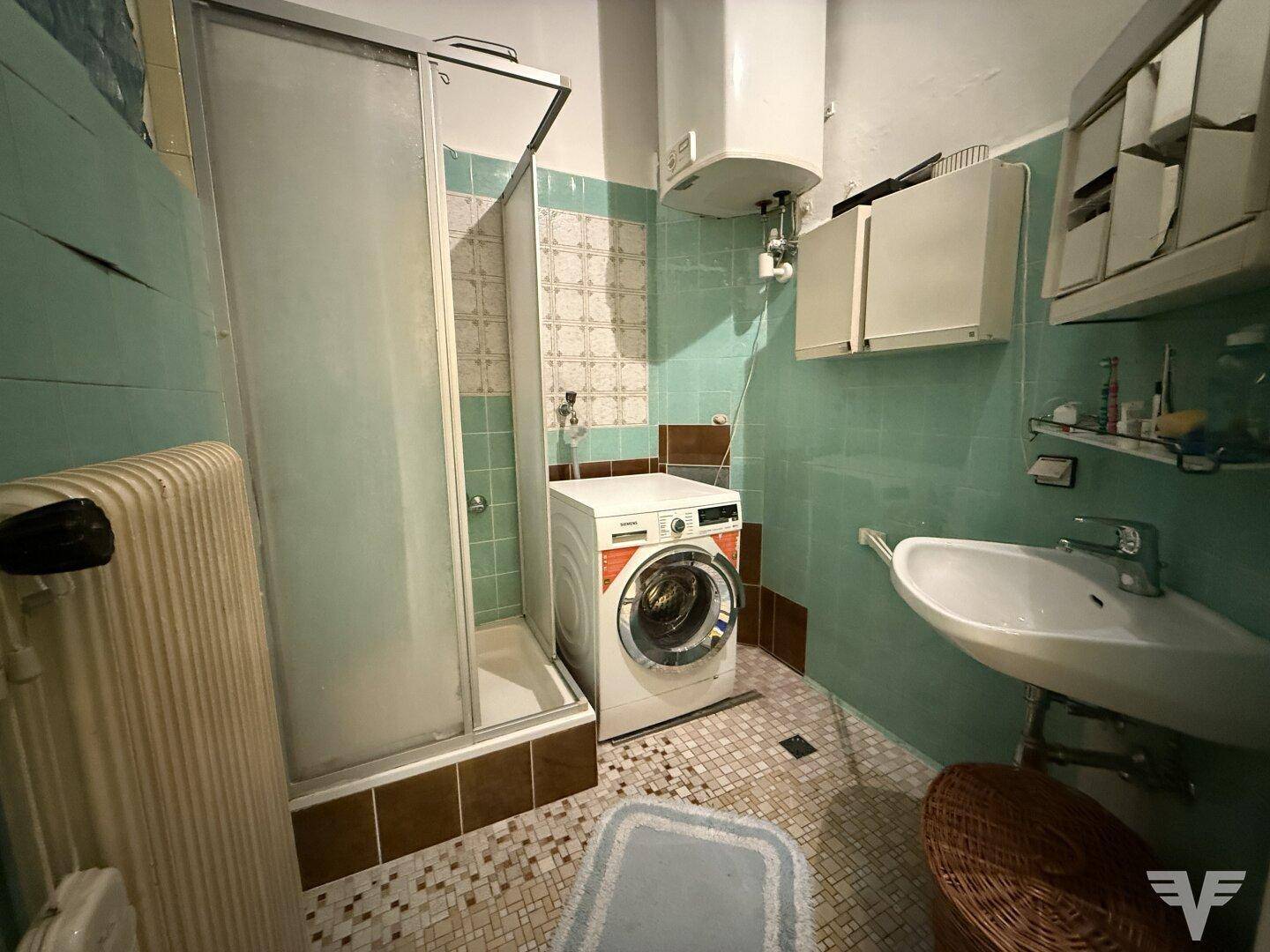 Älteres Badezimmer mit Dusche, Waschmaschine und mintgrünen Fliesen, das eine Modernisierung benötigt.