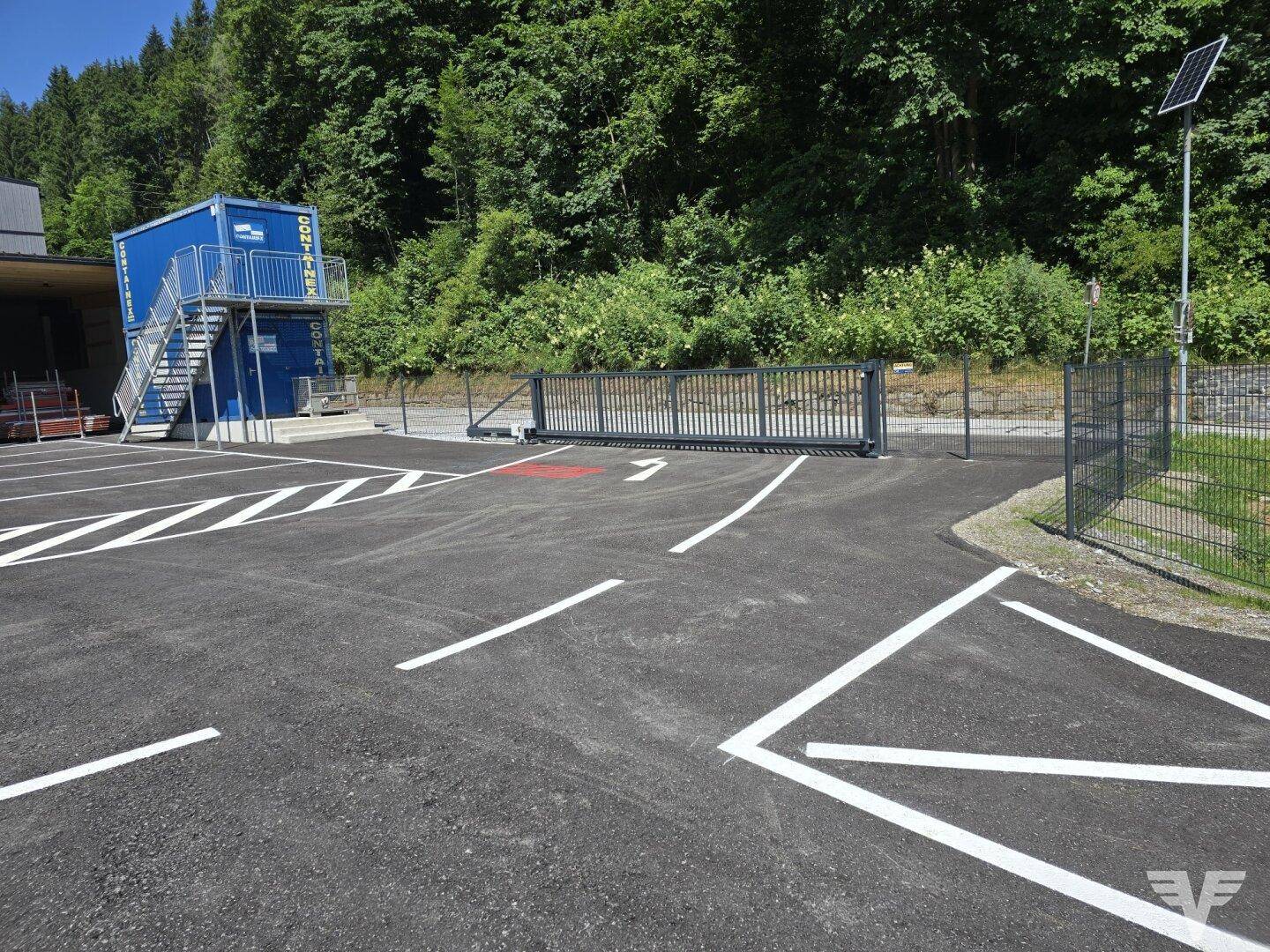 Blick auf den neu angelegten Parkplatz mit einem blauen Containergebäude und einem modernen Zaun.