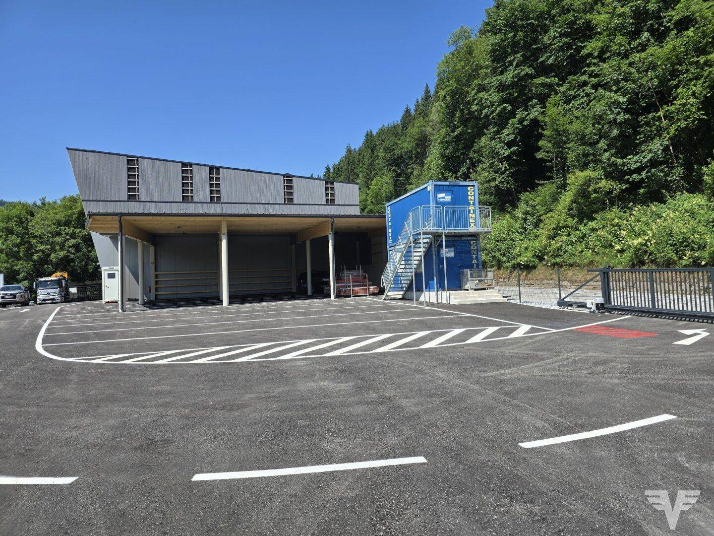 Ansicht des modernen Gewerbegebäudes mit großem Parkplatz und einem blauen Containergebäude im Hintergrund.