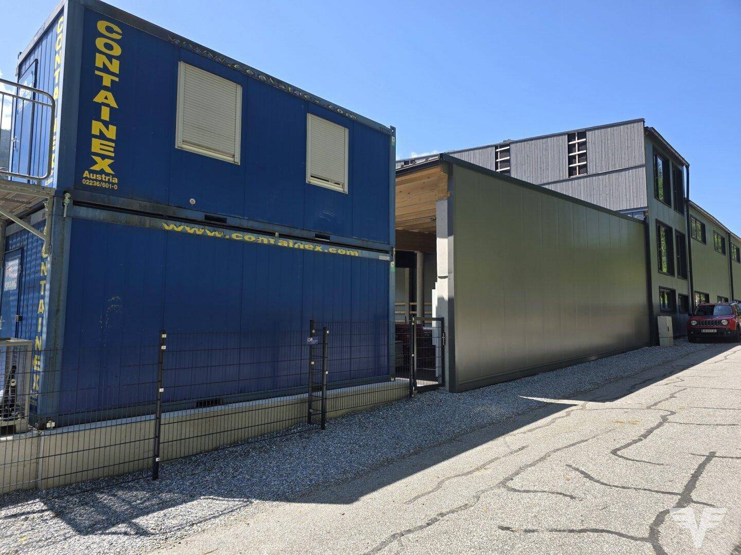 Nahaufnahme des blauen Containergebäudes mit Rollläden und der Seitenfassade des Hauptgebäudes.