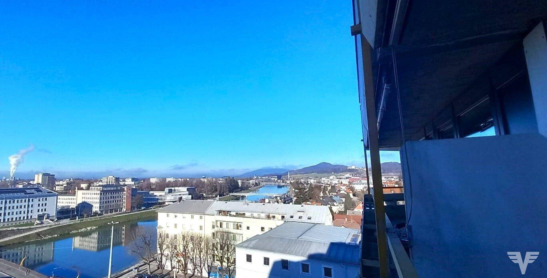 Weitläufiger Stadtblick von oben mit Fluss, Gebäuden und Bergen am Horizont unter blauem Himmel.