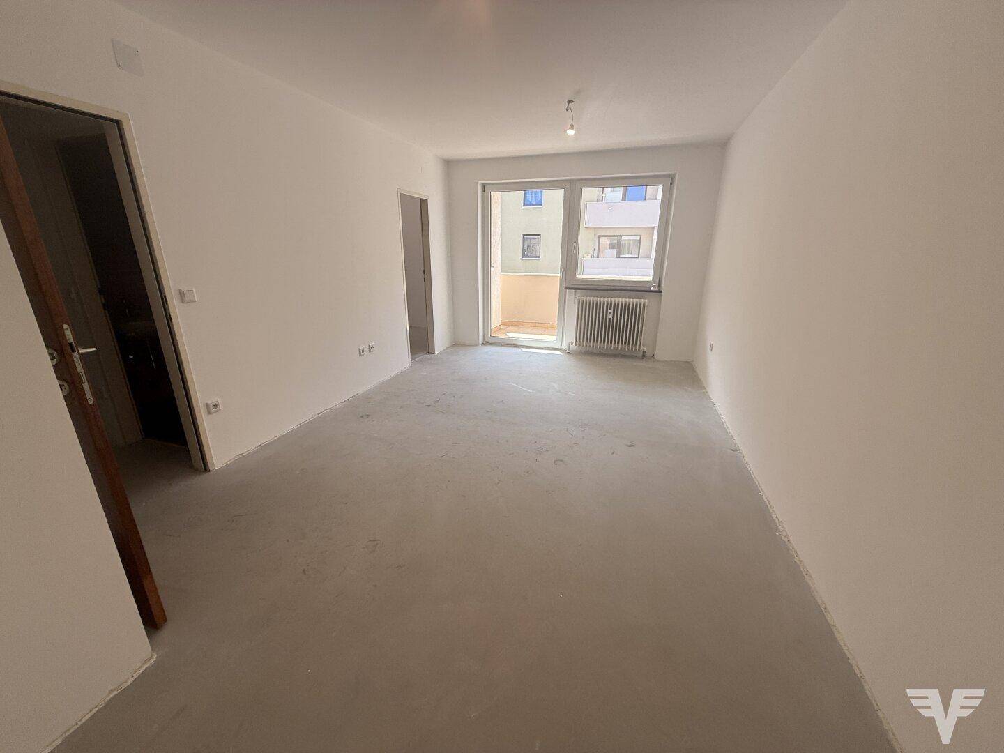 Leerer Raum mit Betonboden und Zugang zu einem Balkon, bereit für den Ausbau.