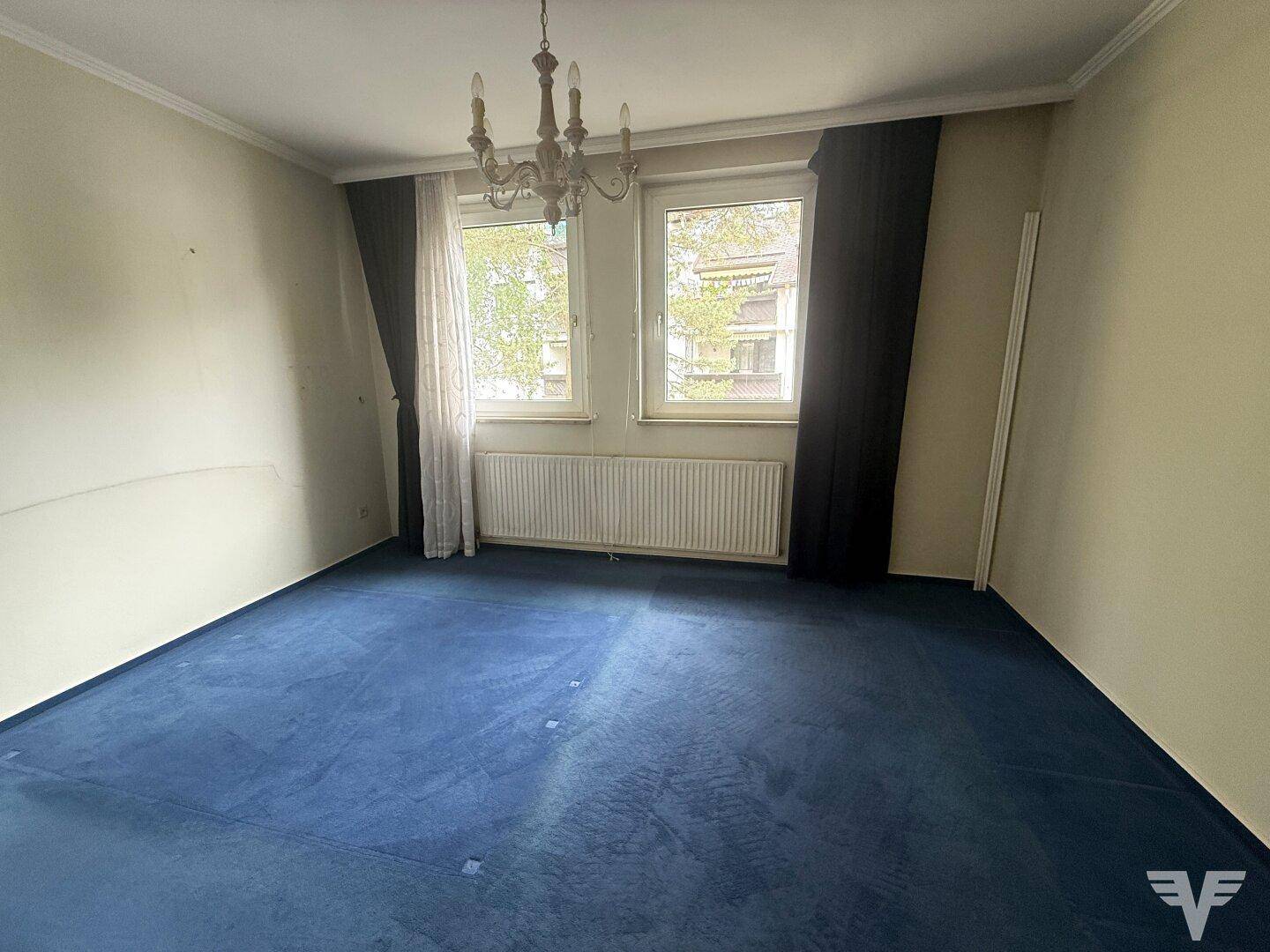 Schlafzimmer mit blauem Teppichboden, großen Fenstern und einem klassischen Kronleuchter.