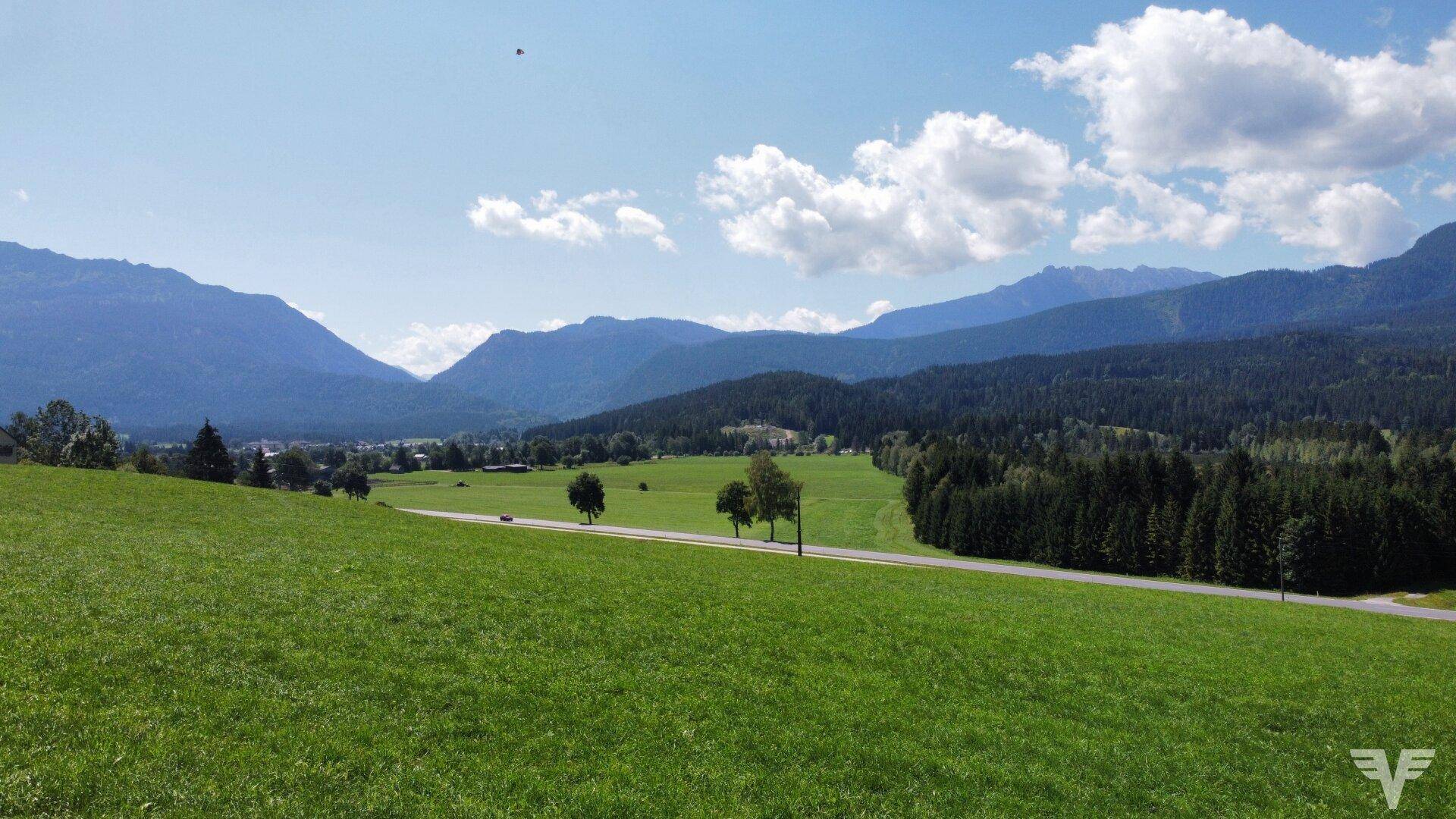 Weitläufige grüne Landschaft mit sanften Hügeln und majestätischen Bergen im Hintergrund unter blauem Himmel.