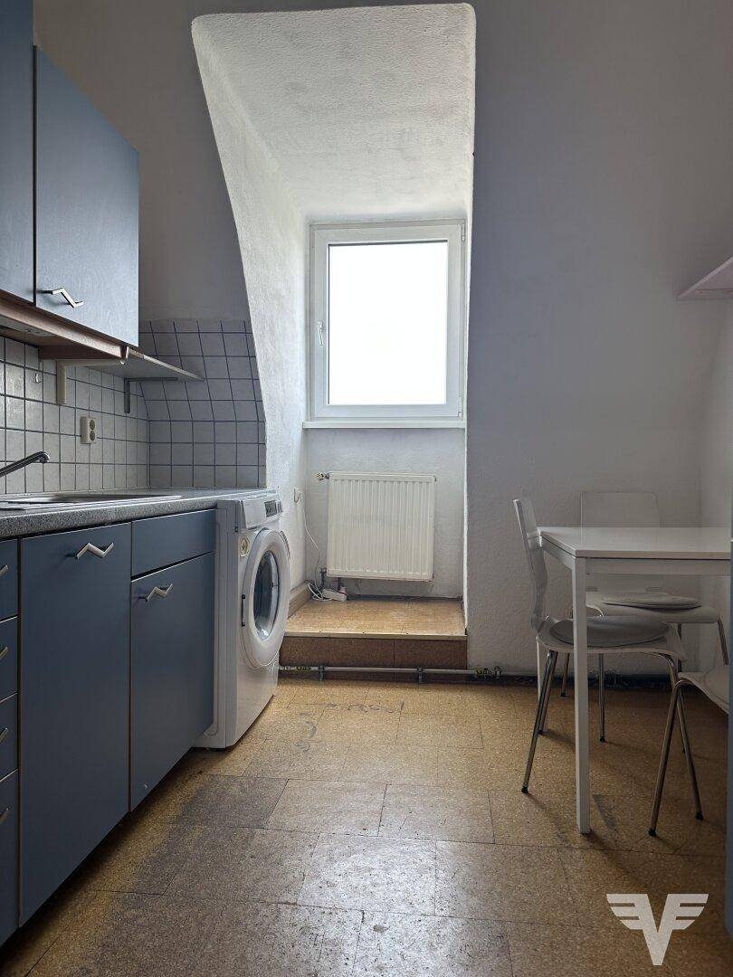 Kleine Küche im Dachgeschoss mit blauen Schränken, Fliesenboden, Waschmaschine und einem Fenster.