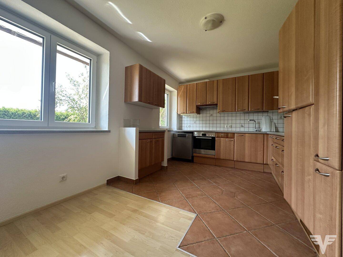 Funktionale Küche mit Holzschränken, Fliesenboden und Blick ins Grüne durch die Fenster.