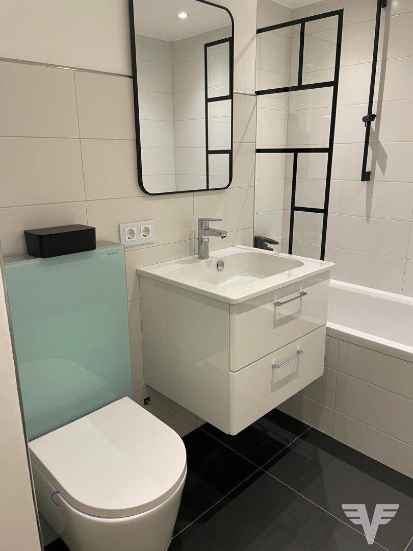 Modernes Badezimmer mit weißem Waschtisch, schwarzen Bodenfliesen und heller Wandverkleidung, inklusive Badewanne und WC.
