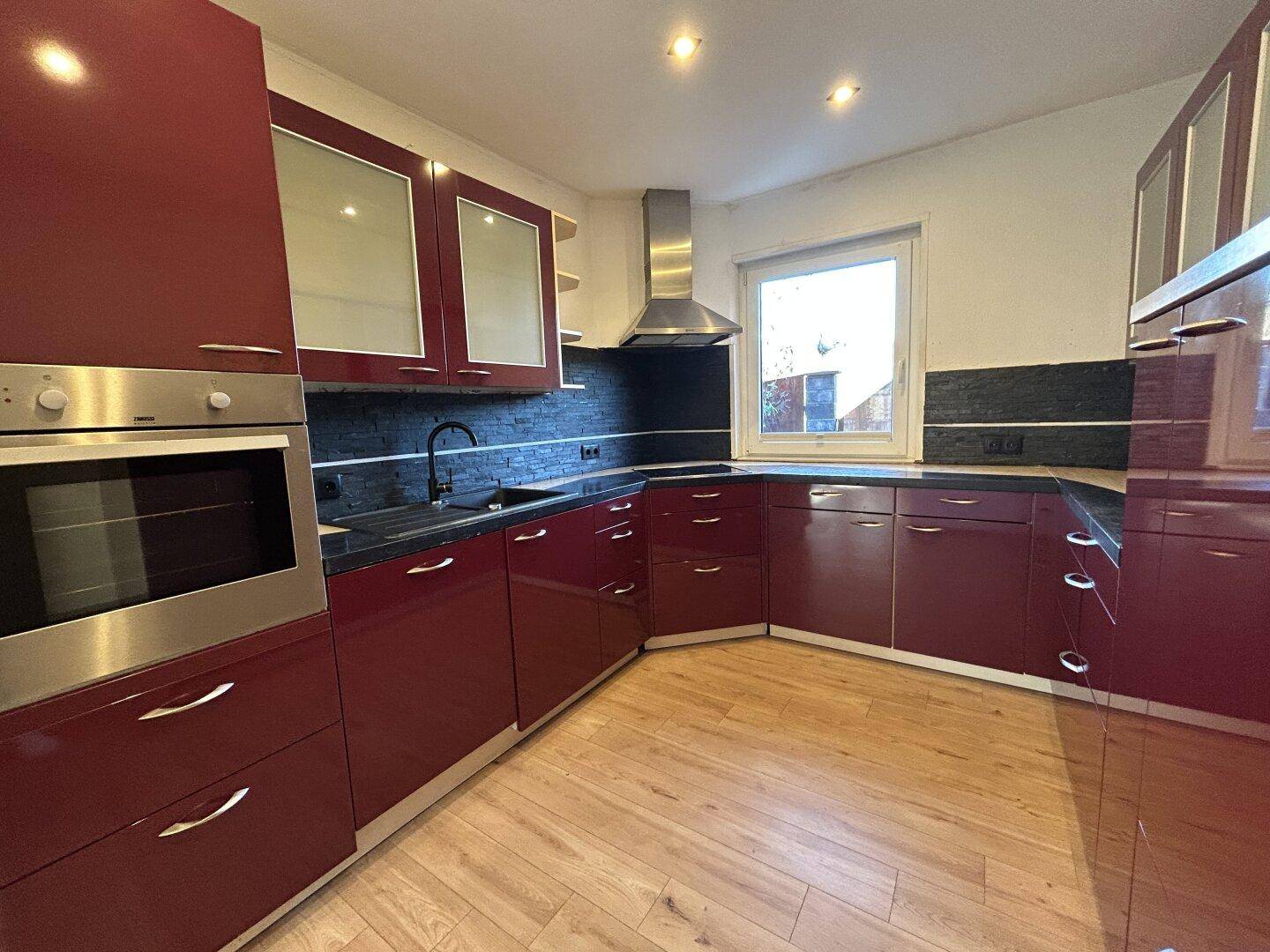 Moderne Küche mit glänzenden weinroten Schränken, schwarzer Arbeitsplatte und integriertem Backofen.