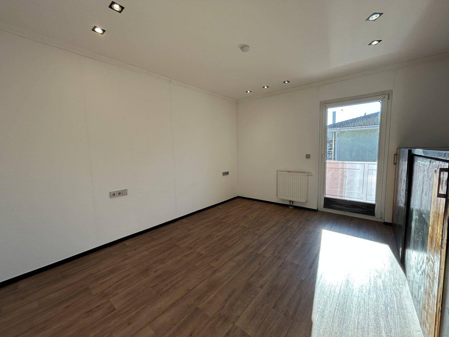 Helles Zimmer mit Laminatboden, Heizkörper und Zugang zu einem kleinen Außenbereich.