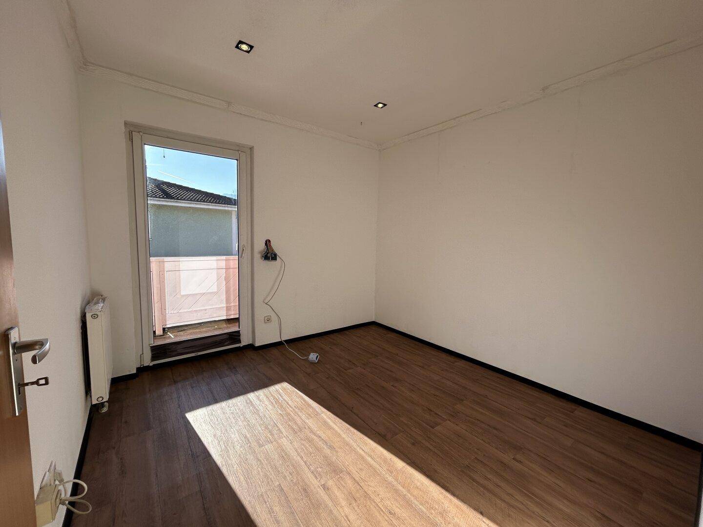 Helles Zimmer mit Laminatboden, Heizkörper und Zugang zu einem kleinen Außenbereich.