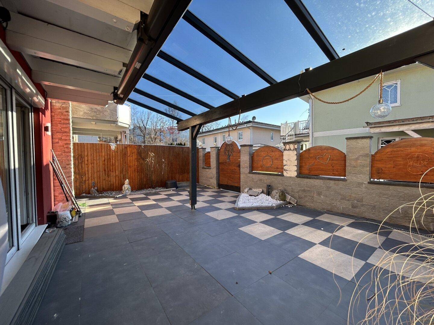 Überdachte Terrasse mit Schachbrettmuster-Fliesen, Holzzaun und Steinmauer im Außenbereich.