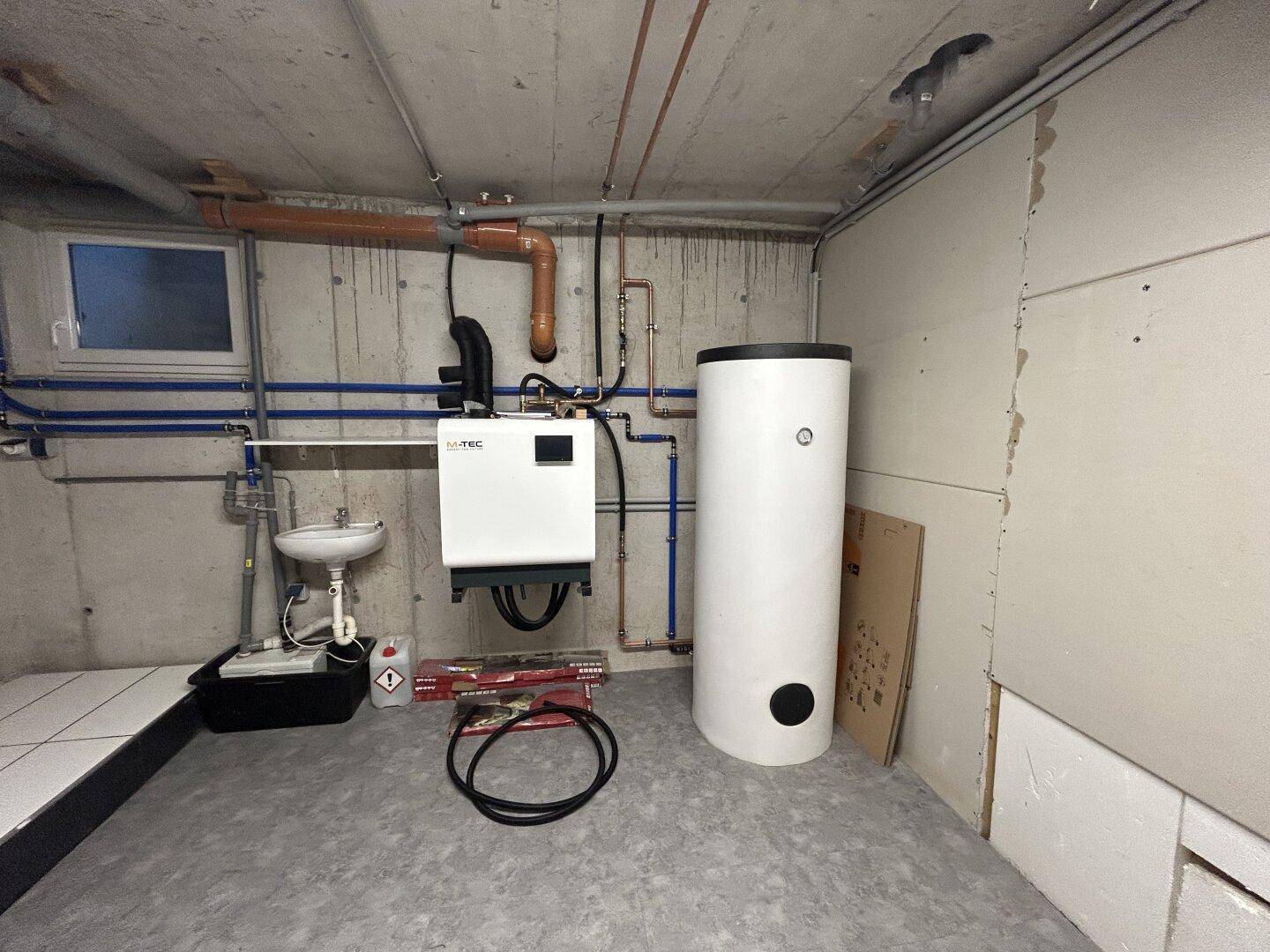 Hauswirtschaftsraum im Keller mit Warmwasserspeicher, sichtbaren Leitungen und kleinem Waschbecken.