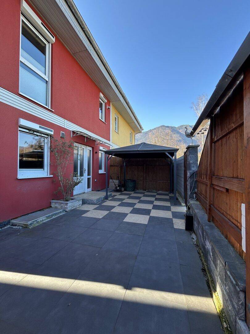Seitenansicht des Hauses mit roter Fassade, gepflastertem Bereich und einem Carport.