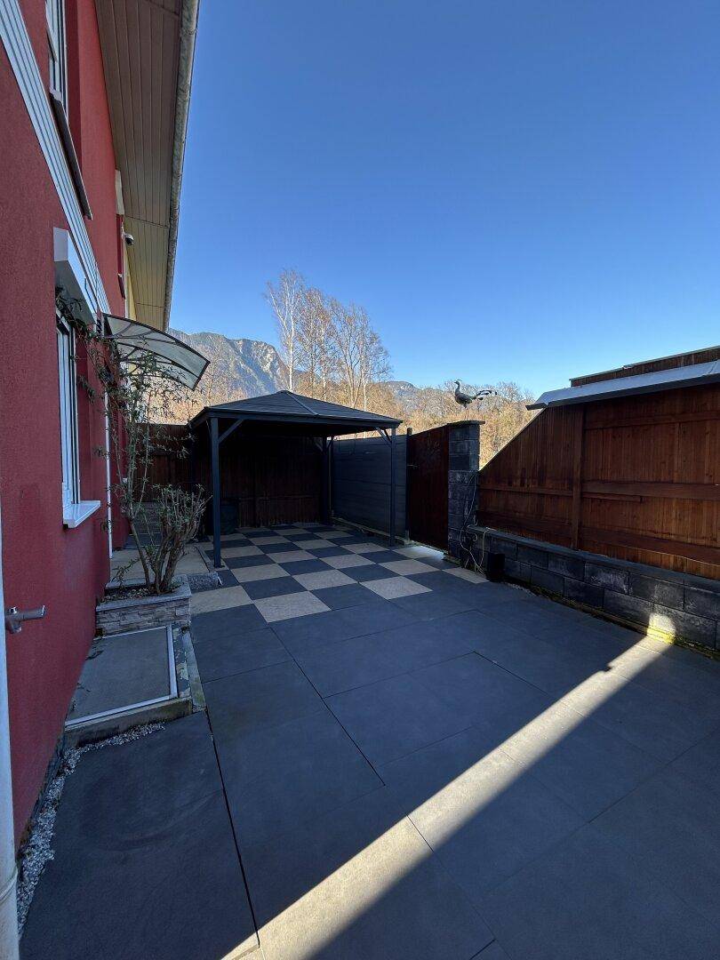 Seitenansicht des Hauses mit roter Fassade, gepflastertem Bereich und einem Carport.