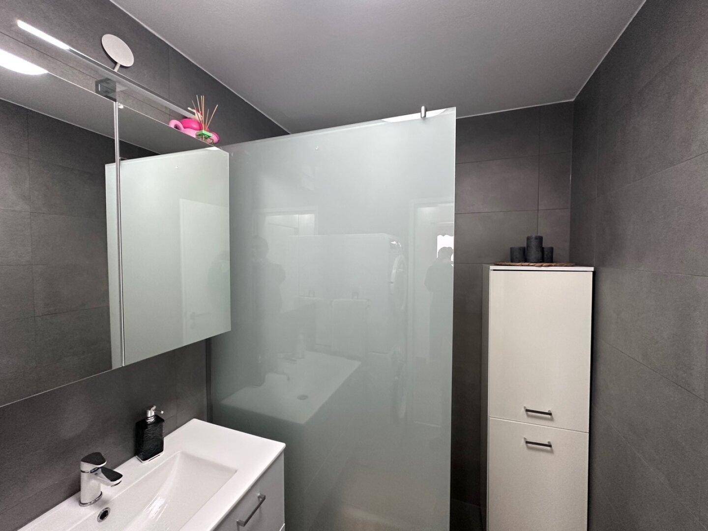 Modernes Badezimmer mit grauen Fliesen, Einzelwaschbecken und einer Duschabtrennung aus Glas.