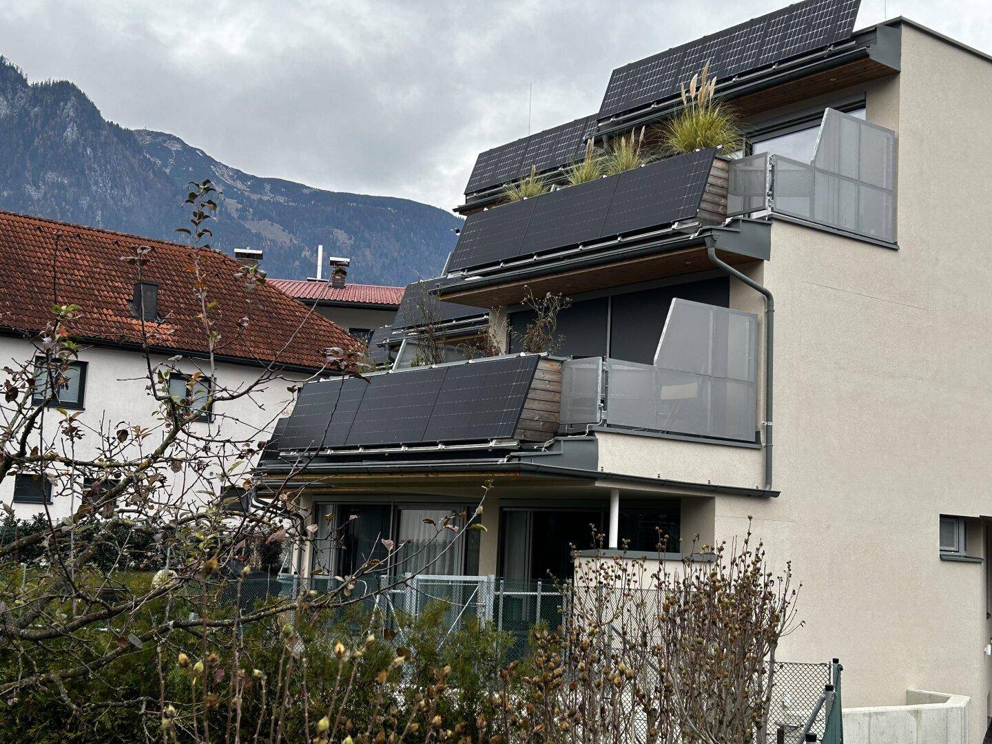 Nahaufnahme der modernen Fassade mit Solaranlagen auf den Balkonen und Bergblick im Hintergrund.