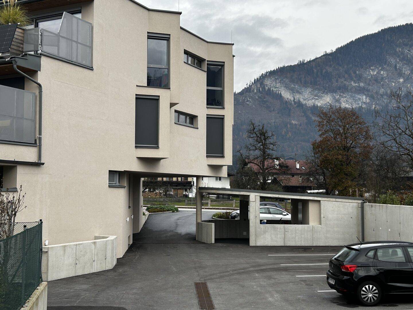 Eingangsbereich und Parkplätze des modernen Gebäudes mit Bergblick unter bewölktem Himmel.