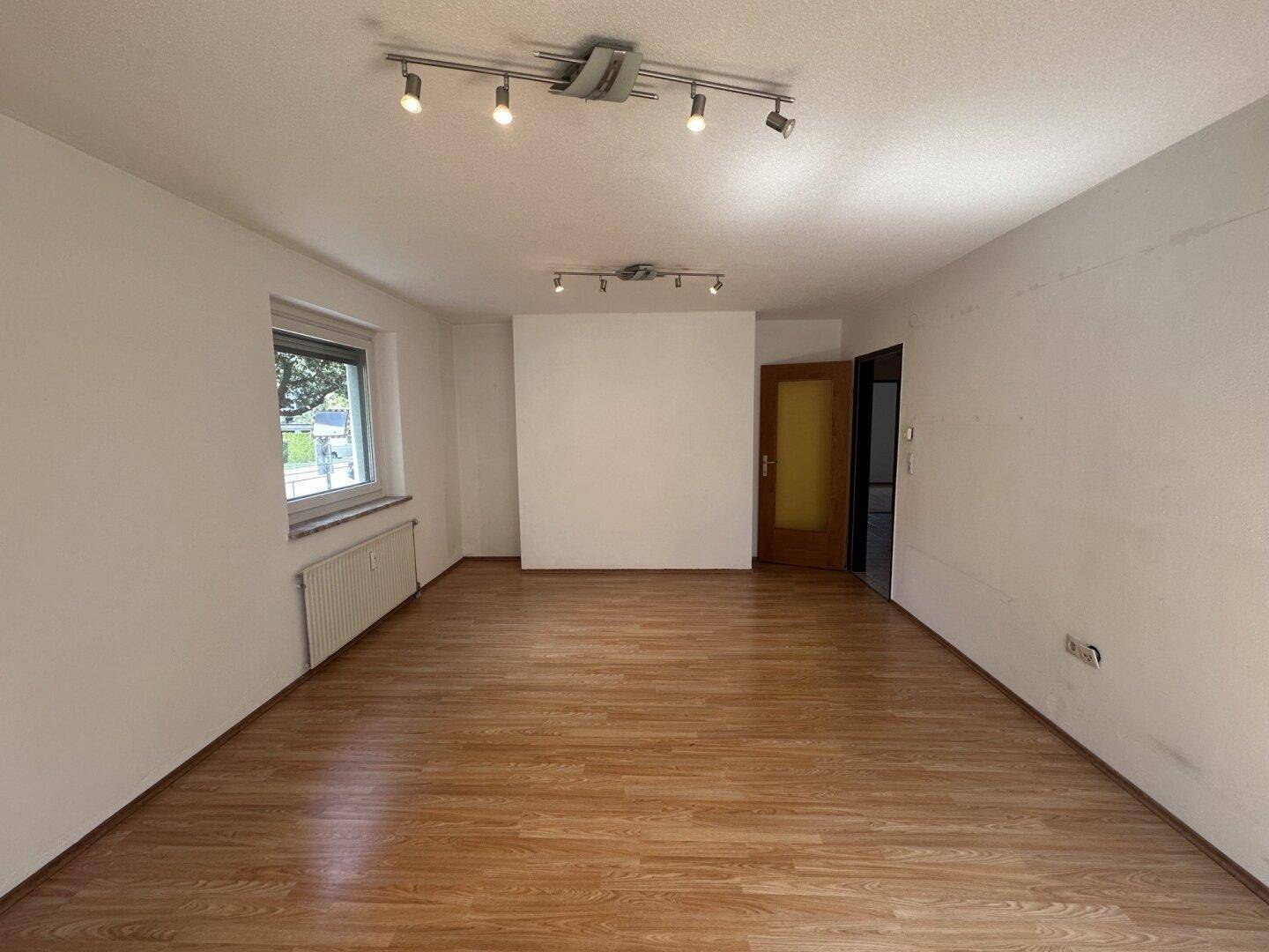 Leeres Wohnzimmer mit Laminatboden, weißen Wänden und einer Holztür.
