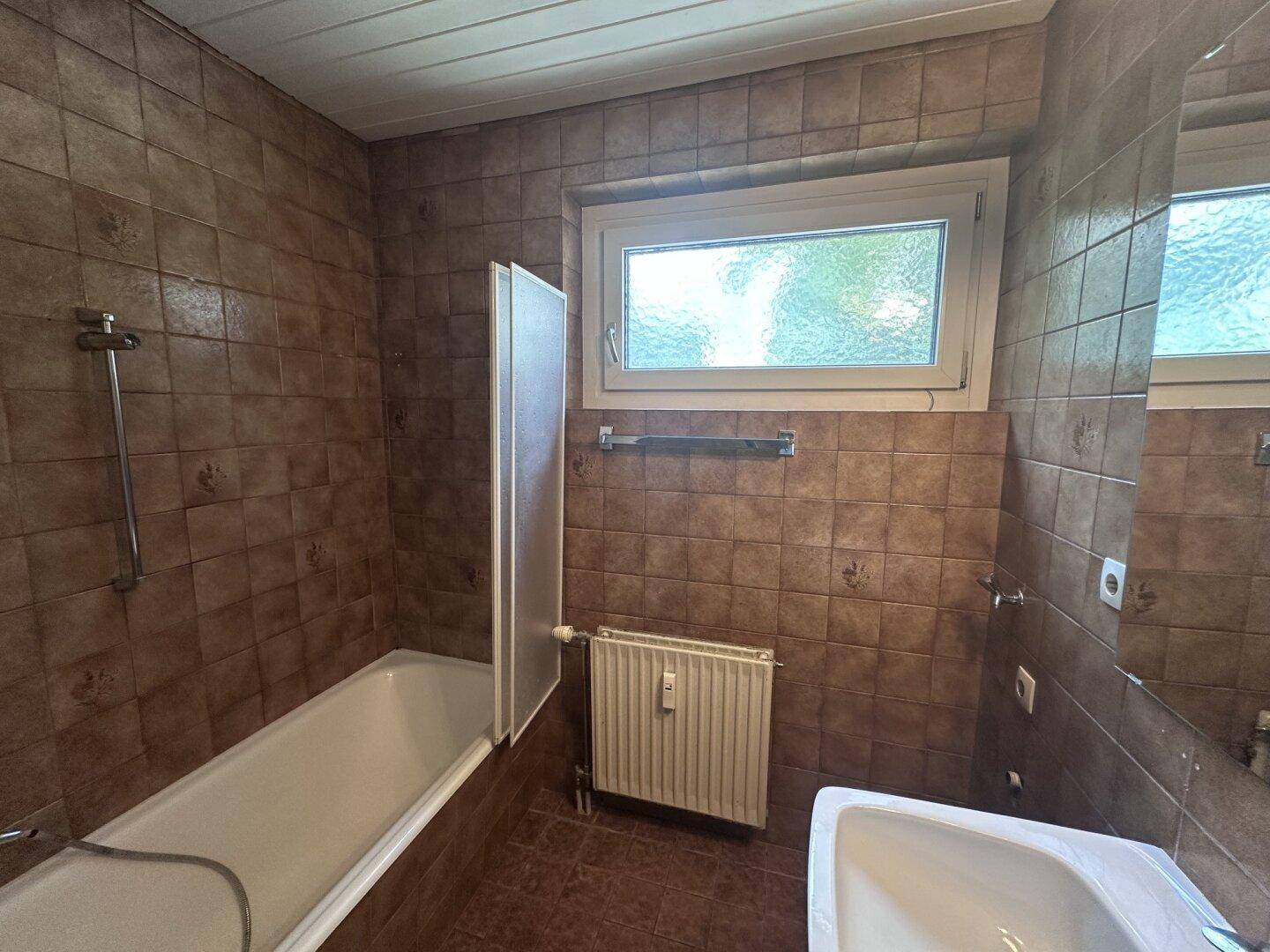 Badezimmer mit braunen Fliesen, Badewanne und Fenster für natürliche Belüftung.