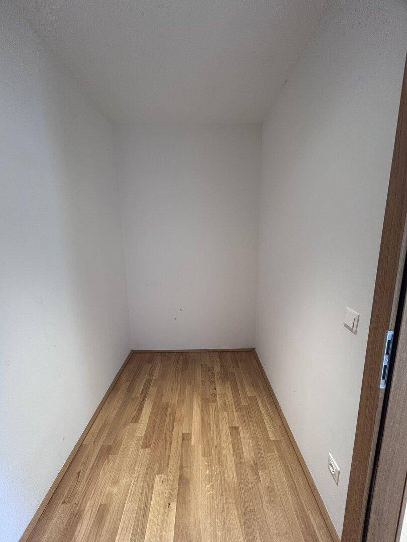 Leerer Abstellraum mit hellem Holzparkettboden und weißen Wänden in neuwertigem Zustand.