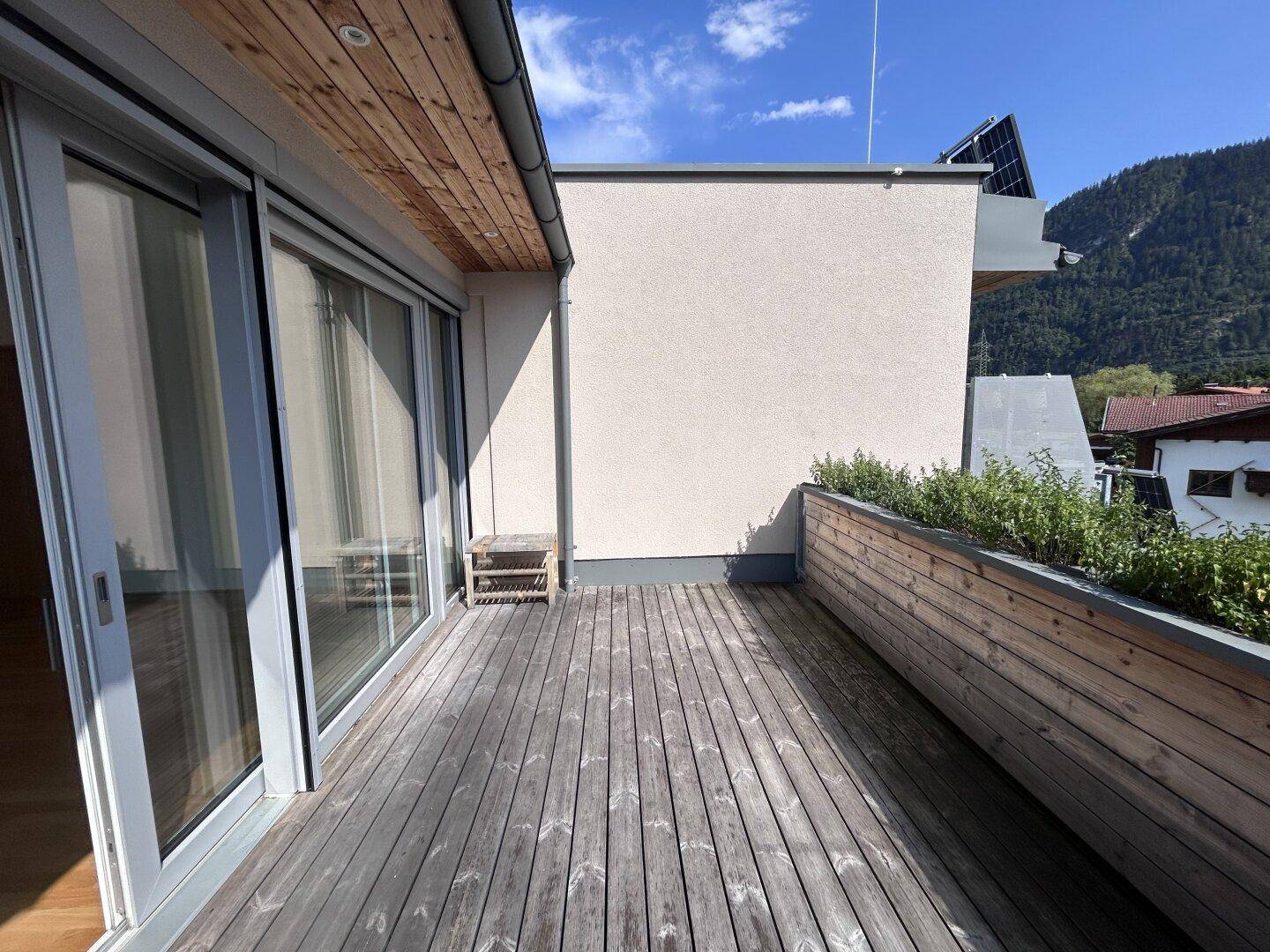 Weitläufige Holzterrasse entlang der Gebäudefassade mit integriertem Pflanzenbereich.
