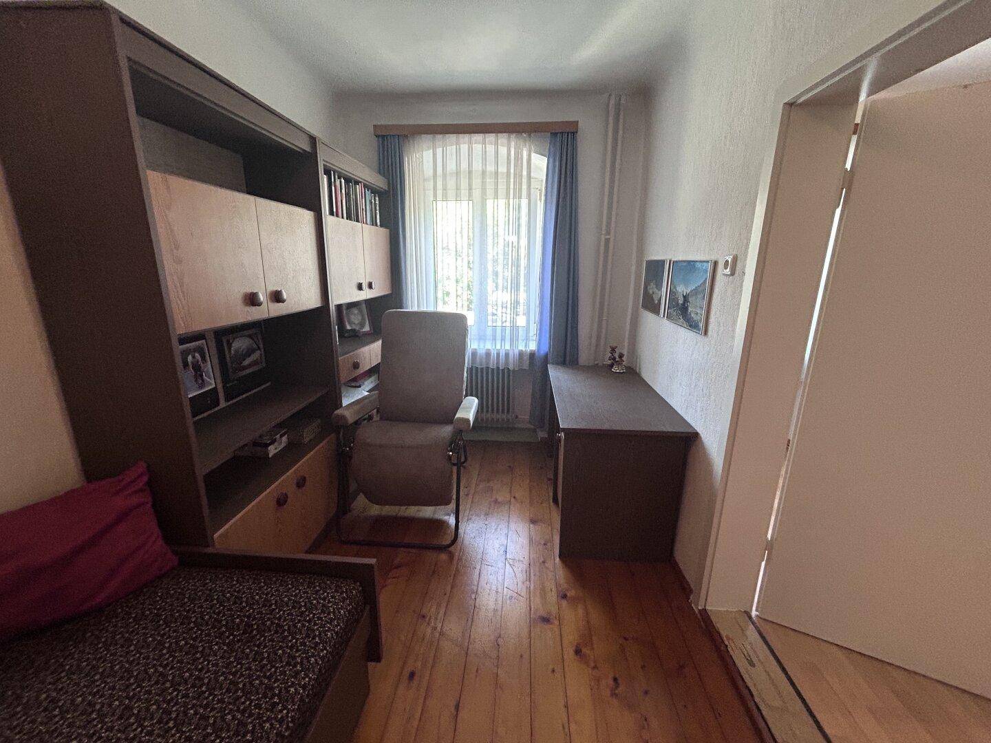 Kleines Arbeitszimmer mit Holzfußboden, Schreibtisch und einem Sessel am Fenster.