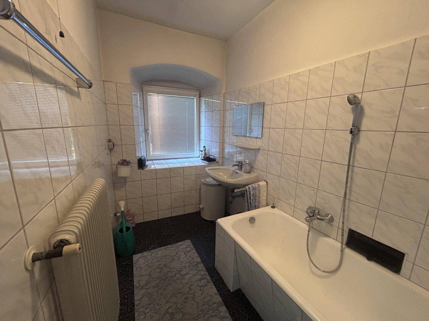 Badezimmer mit Badewanne, Waschbecken und weißen Fliesen an den Wänden, hell durch das Fenster.