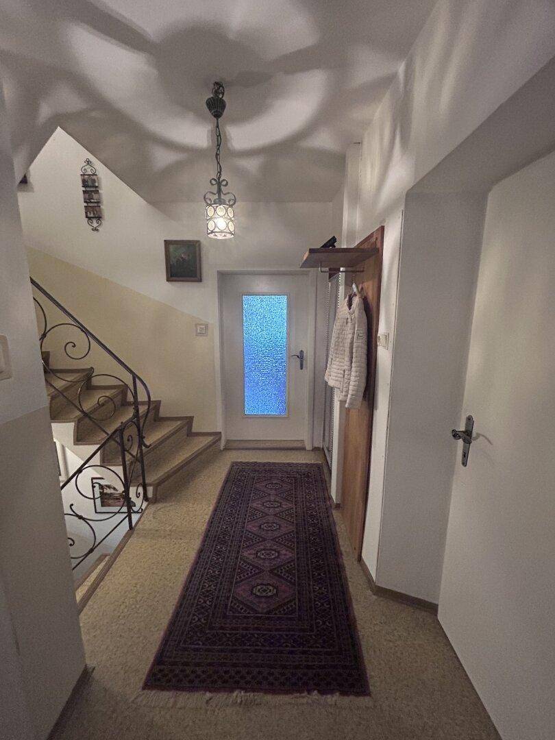 Eingangsbereich mit Treppenaufgang, Teppichläufer und einer Garderobe, hell durch die Glastür.